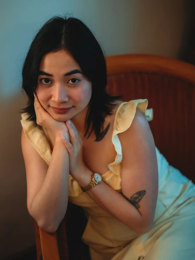A woman in a yellow dress sits in a rattan chair, her chin resting on her hands, a gold watch on her wrist, in warm light against a teal wall.