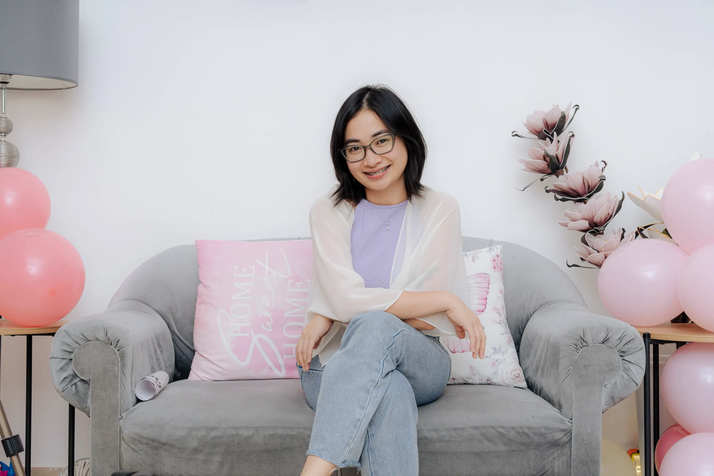 A smiling woman in glasses sits on a grey sofa with pink cushions, pink balloons around her.