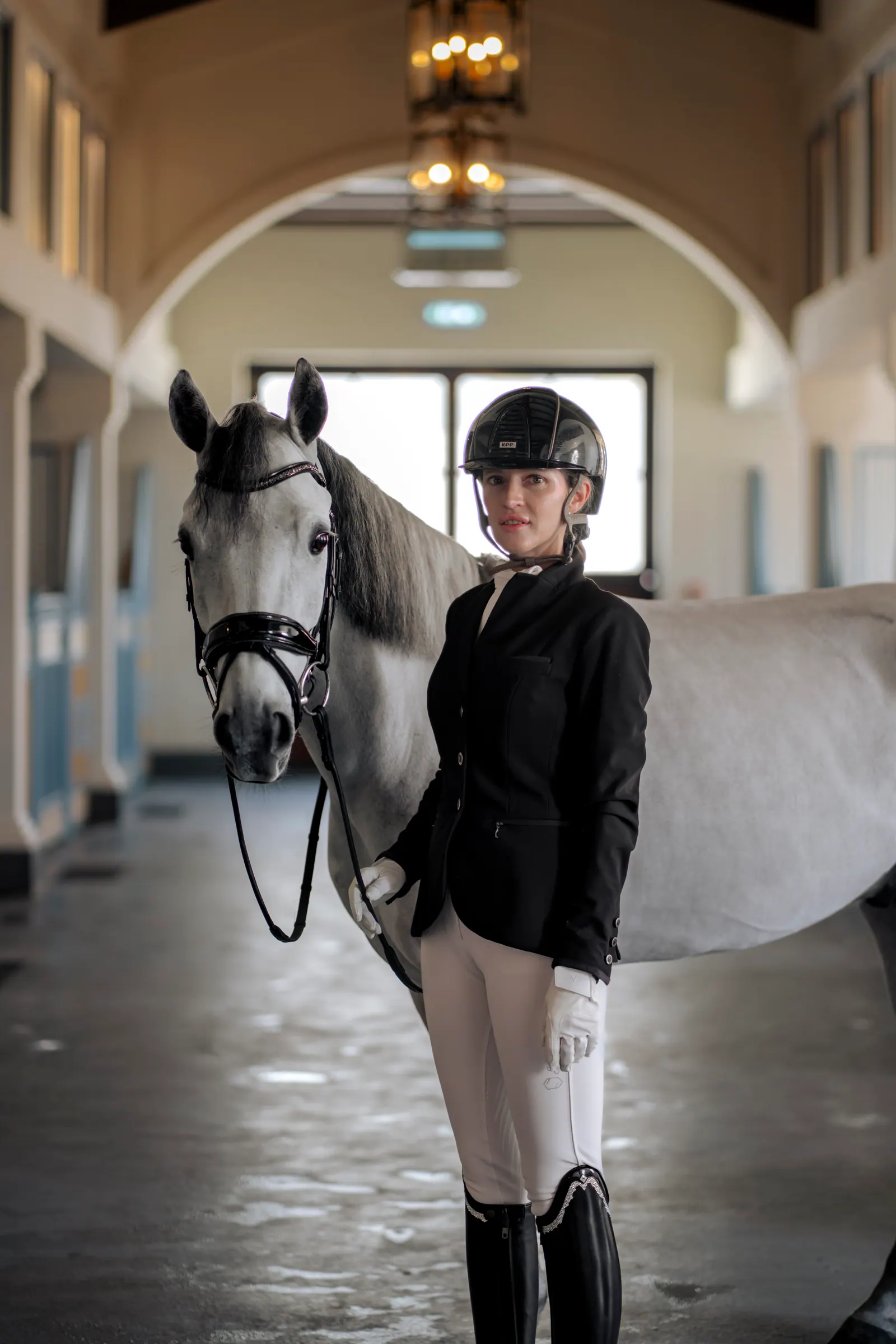A rider in a black show jacket, helmet and white gloves stands beside a grey horse in its bridle.