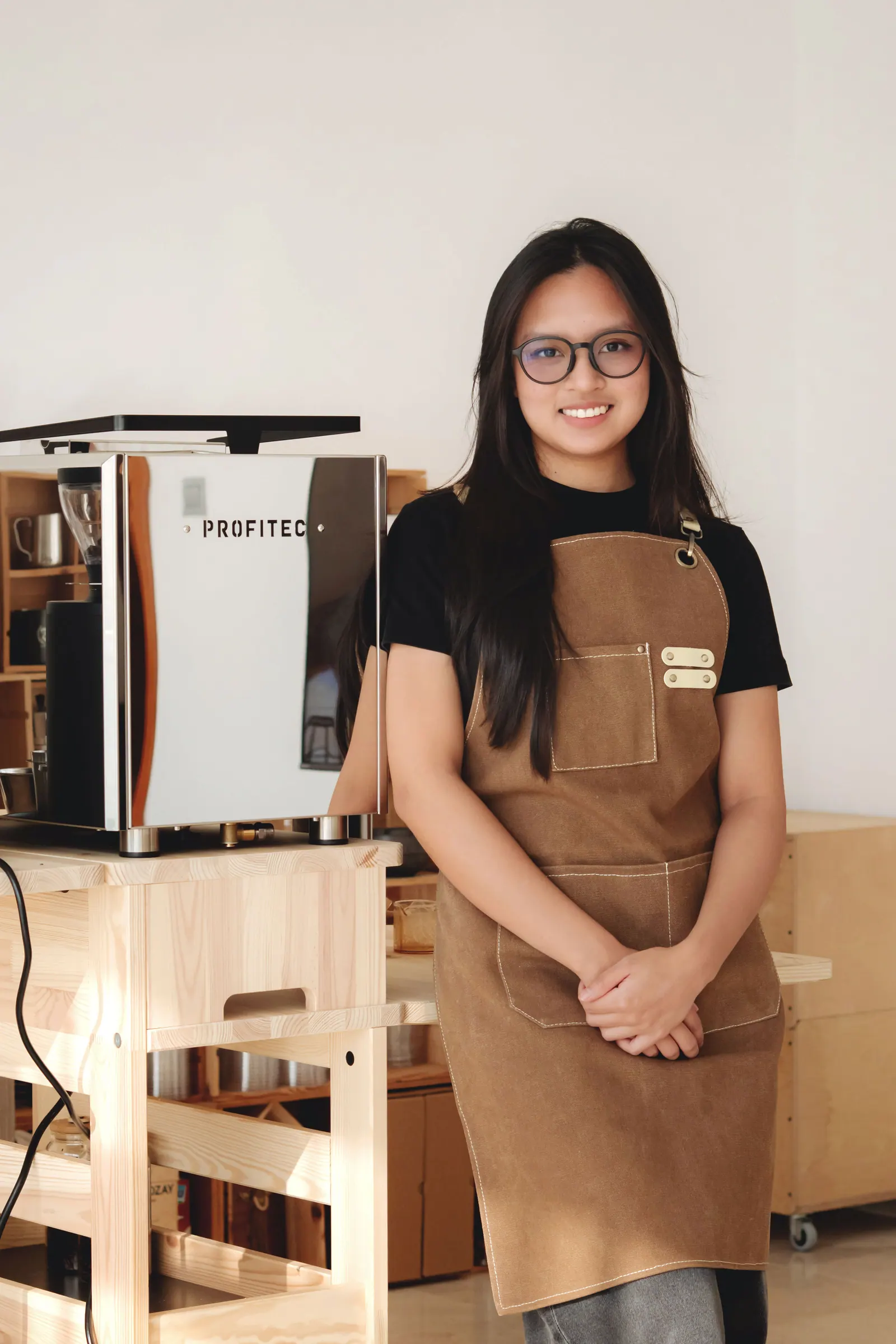 A smiling barista in glasses and a brown apron stands beside an espresso machine.