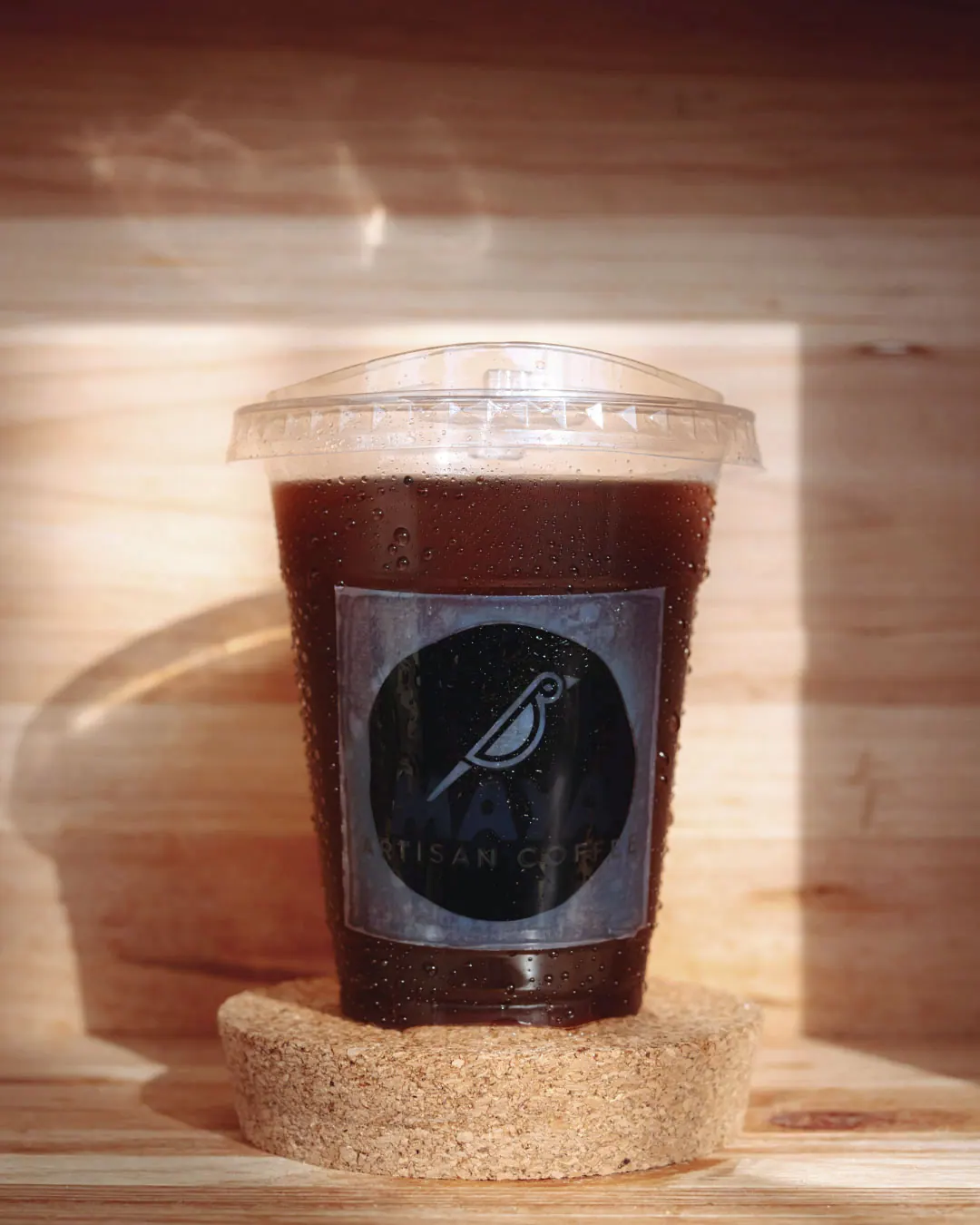 An iced black coffee in a Maya Artisan Coffee cup on a cork coaster inside a wooden crate.