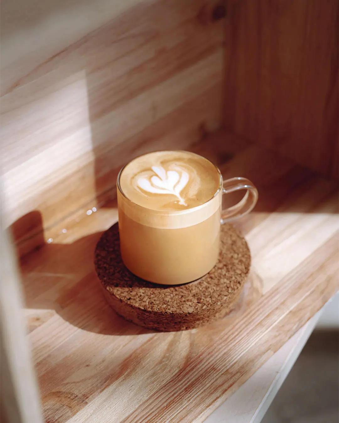 A cappuccino with heart latte art in a glass mug on a cork coaster.