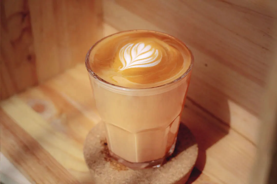 A latte with leaf latte art in a glass tumbler on a cork coaster, lit by warm side light.