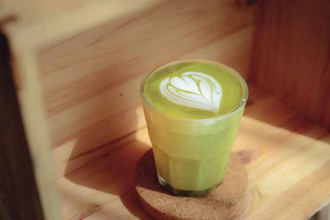 A matcha latte with heart latte art in a glass on a cork coaster.