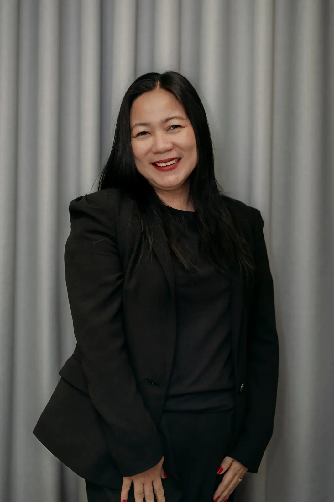A smiling adult with long dark hair in a black blazer stands at an angle before a grey curtain.