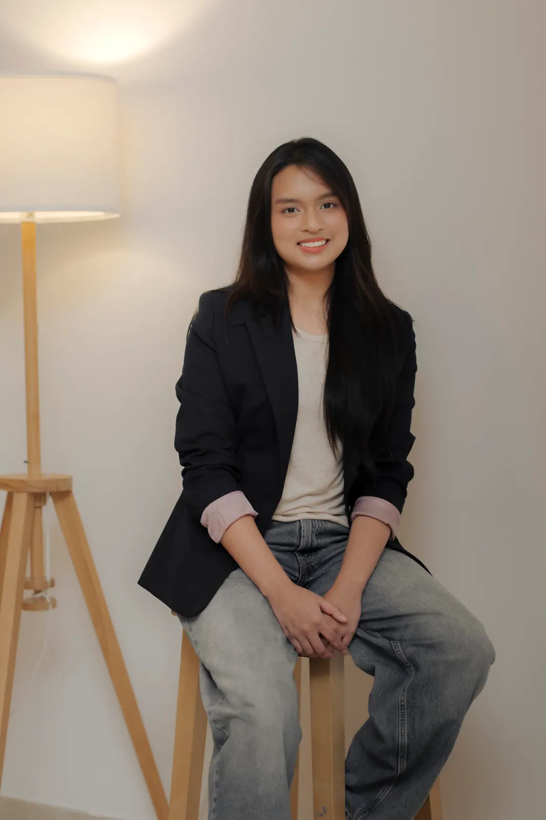 A young adult with long dark hair in a black blazer and jeans sits on a stool beside a floor lamp.
