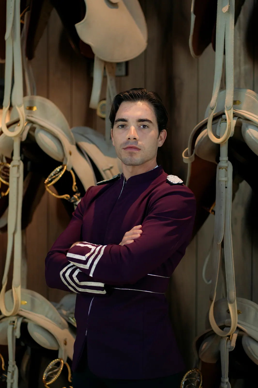 A rider in a plum uniform with white braid stands with arms crossed in a tack room lined with saddles.