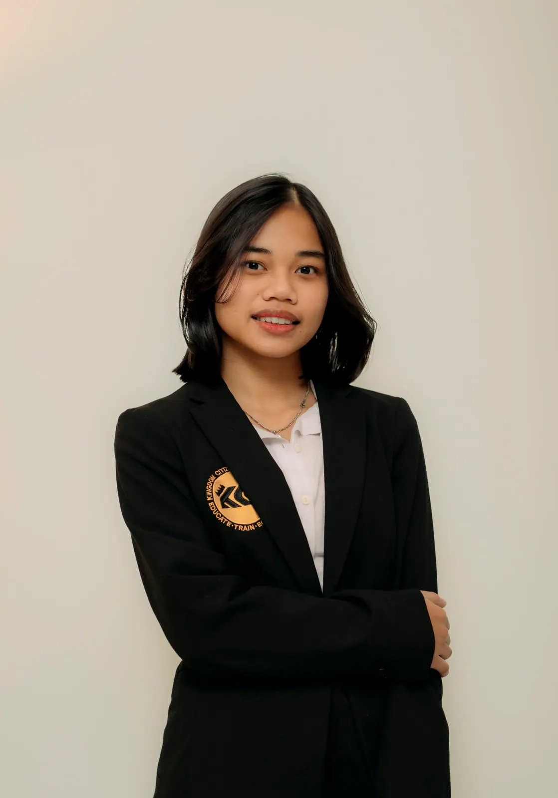 A teenager with shoulder-length hair in a black crested blazer stands with arms folded against a cream backdrop.