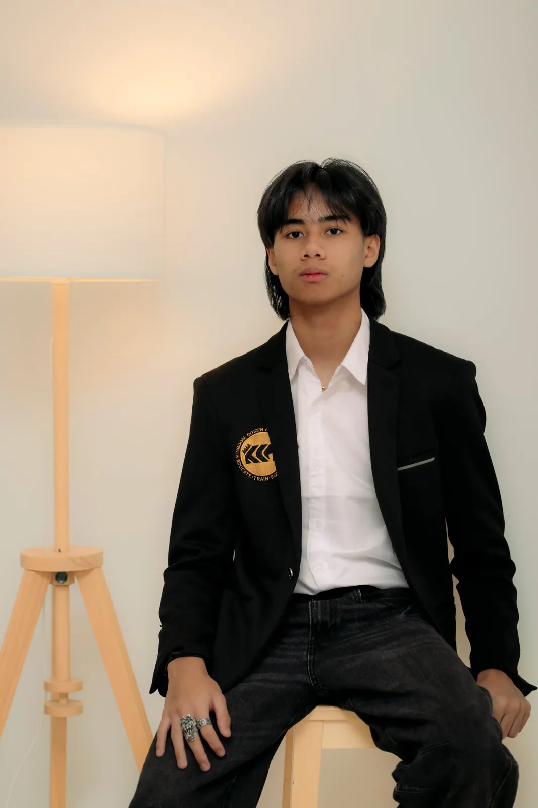 A teenager with chin-length hair sits on a wooden stool in a black crested blazer and dark jeans.